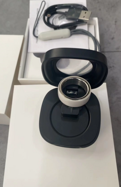 A black circular magnetic charging dock or stand holding a small cylindrical device, shown with its white box and a USB charging cable in the background on a gray floor.