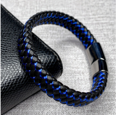 A braided leather bracelet with a vibrant blue center stripe and a polished silver-tone metal clasp, displayed on a textured black leather surface against a gray fabric background.
