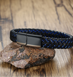 A braided black and blue leather bracelet with a matte black magnetic clasp, resting on a textured brown rock over a wooden surface.