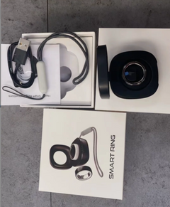 Smart ring kit with a black smart ring, charging cable, and accessories displayed on top of their white retail packaging boxes, set on a grey tiled floor.