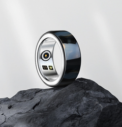 A smart ring with a polished silver outer band and a glossy black inner face, standing upright on a dark textured rock against a light gray background.