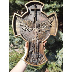 Wooden cross with angel and shield design held against a green foliage background