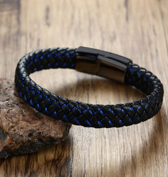 A braided black leather bracelet with blue accent stitching and a black magnetic clasp, resting on a textured brown rock over a wooden surface.