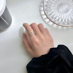 A close-up of a person's hand wearing three delicate silver rings with small clear stones, styled on a white tabletop with a glass plate and a cup in the background.